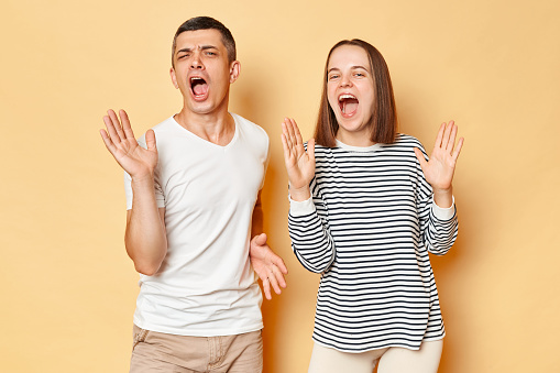 Positive optimistic young couple wife and husband wearing casual attires standing isolated over beige background dancing and singing songs having fun being in good mood.