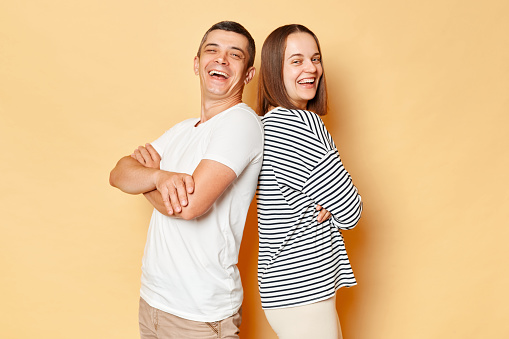 Happy cheerful young couple wife and husband wearing casual attires isolated over beige background standing back to back looking at cmaera and laughing having fun together.