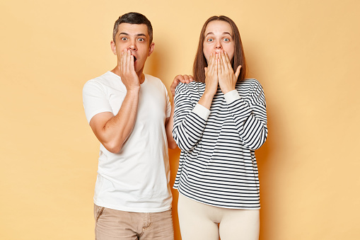 Shocked young couple wife and husband wearing casual attires standing isolated over beige background looking at something scary covering mouths with palms being afraid.