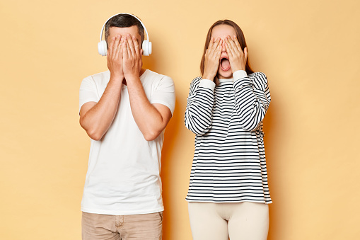 Scared young couple wife and husband wearing casual attires standing isolated over beige background covering eyes with hands screamign feeling fear.