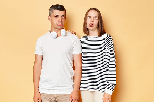 Funny childish young couple wife and husband wearing casual attires standing isolated over beige background frimacing showing tongue out and pout lips playful behavior.