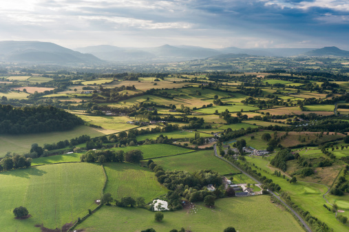 Rays of sunlight illuminating the picturesque patchwork quilt landscape of green fields, farms and misty mountains under panoramic skies from high in a hot air balloon. ProPhoto RGB profile for maximum color fidelity and gamut.