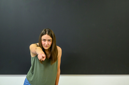 Angry looking female instructor standing at the blackboard in a classroom pointing at camera