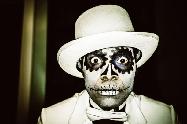 Man with typical Mexican-style Day of the Dead (Dia de los Muertos), sugar skull make-up, dressed in white suit and hat. Dark, Surreal Portrait.