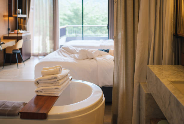 Fresh bathroom textile in cozy hotel room with bright interior. Selective focus on stack of dry and soft beige towels on comfortable bed with bedding and cushions.