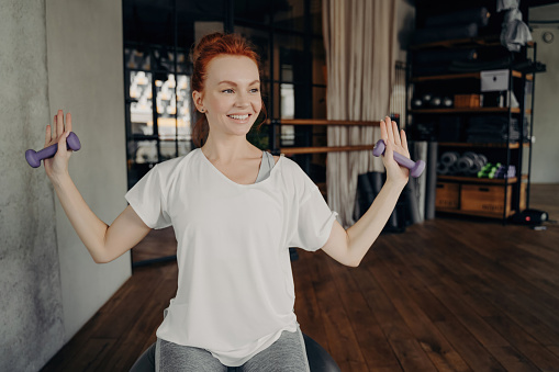 Happy European redhead woman in active wear sits on silver fitball, raising arms with dumbbells in fitness studio. Positive workout vibes!