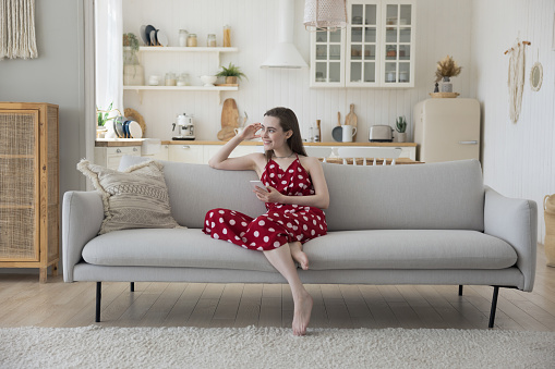 Happy thoughtful pretty adult girl with smartphone sitting on comfortable home sofa, looking away, smiling, enjoying positive thoughts, plans, thinking on text message, Internet shopping