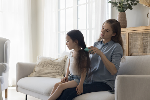 Caring young mom brushing black long hair of cute little girl kid, helping child with girlish beauty care, enjoying childcare, motherhood, maternity leave, family leisure at home