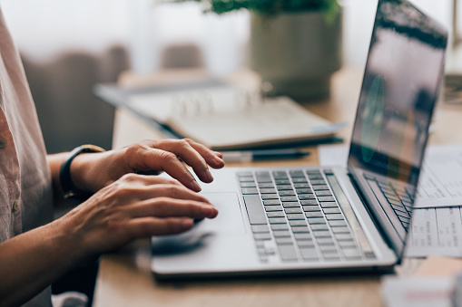 A side view of an unrecognizable Caucasian entrepreneur typing on her laptop. She is sitting at her office desk.