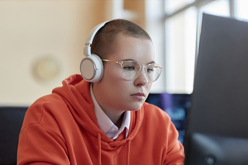 Young businesswoman in eyeglasses, red hoodie and headphones sitting in front of computer screen with data in office and analyzing it