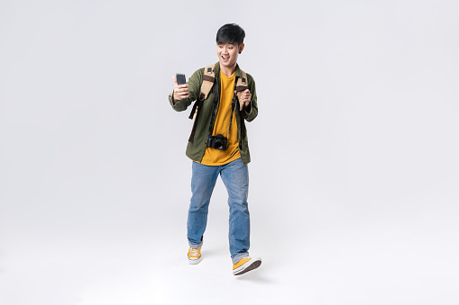 Full length happy Asian man traveller walking with using smartphone for check in or shopping online. Isolated in studio white background. Technology communication, Internet.