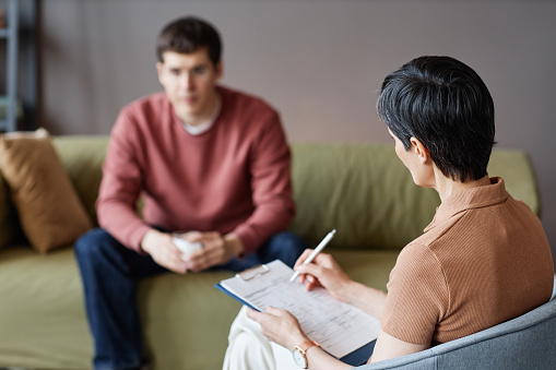 Rear view of mature psychologist making notes in card during conversation with patient