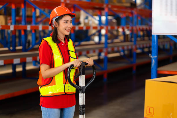 istock Warehouse female staff worker wearing hardhat helmet standing by goods shelf working pulling a pallet truck in large warehouse factory. Asian engineer woman 1564430311