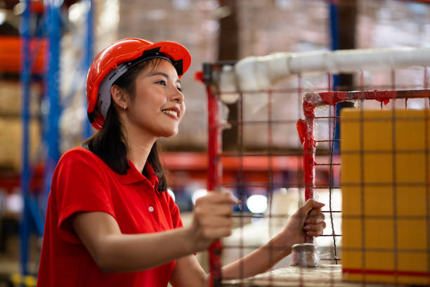istock Warehouse female staff worker wearing hardhat helmet standing by goods shelf working pulling a pallet truck in large warehouse factory. Asian engineer woman 1564430316