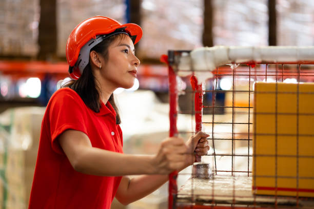 istock Warehouse female staff worker wearing hardhat helmet standing by goods shelf working pulling a pallet truck in large warehouse factory. Asian engineer woman 1564430825