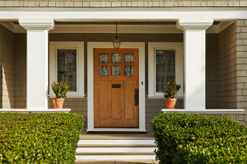 A concrete walkway bordered with hedged shrubs leads to the front door of a home. There are windows on either side of the door. Horizontal shot.