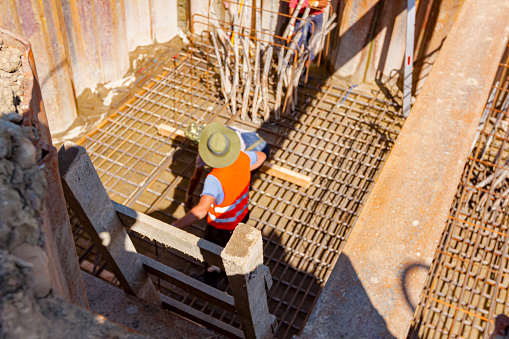 Shot from above of the ladder for safe descent of workers into trench surrounded by metal piles. Installing basic profile made of reinforcing rods for bridge foundation, teamwork on construction site.