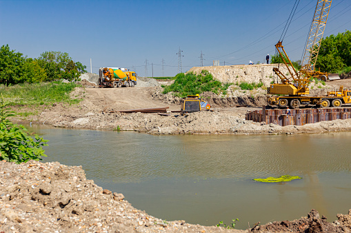 Across the water is yellow obsolete mobile crane with pneumatic hammer. Installed metal piles along river bank built in for bridge foundation at the construction site.