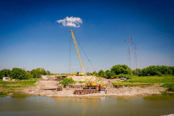 istock Over river is old hoist with pneumatic hammer on building site. Bridge foundation made of metal piles on coast 1564607722