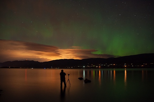 A silhouette of a photographer with a camera on a tripod under the Northern Lights on Okanagan Lake