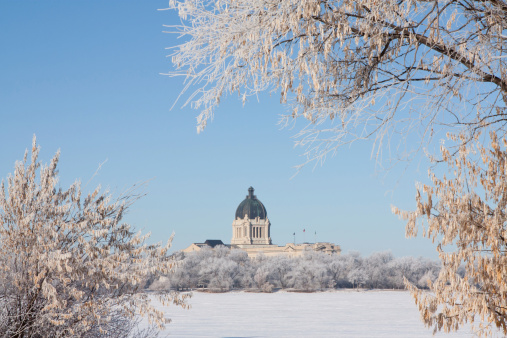 The Saskatchewan Legislative Building in Regina, Saskatchewan, Canada is framed by frost covered trees in winter.  Snow-covered Wascana Lake is in between.
