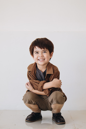 A portrait of a boy wearing a brown polite shirt squatting and crossing his arms.