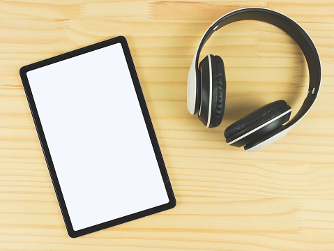 Top view or flat lay of digital tablet with white blank screen and headphones on wooden table background.