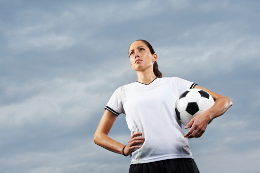 A footballer holds a football under her arm and looks towards the audience.