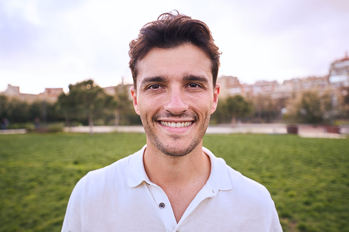 Portrait of attractive smiling young man looking at camera in park. Caucasian male handsome model poses cheerful for photo outdoors. Unfocused background with copy space. Face positive expression.