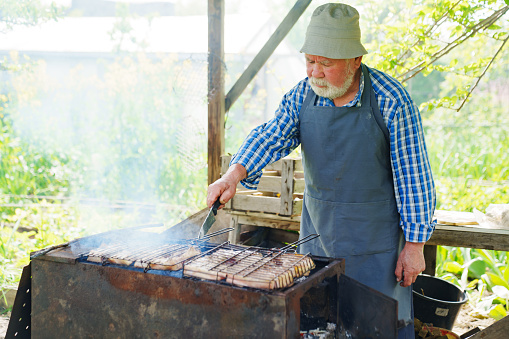 an elderly man with a beard in work clothes cooks meat steaks on the grill over an open fire. Picnic menu.