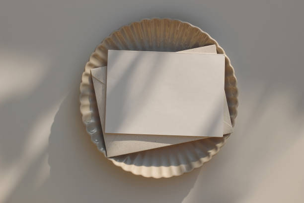 istock Summer minimal wedding stationery. Blank horizontal greeting card mockup with beige envelope, invitation. Ceramic scallop dinner plate in sunlight. Table background with shadows overlay. Flatlay, top 1564947578
