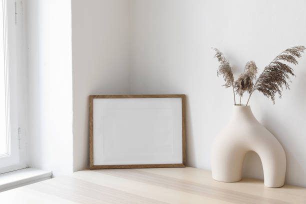 istock Dry grass, reed plant bouquet in modern ceramic vase. Empty photo frame mockup against white wall on wooden table near window. Minimalistic Scandinavian style, elegant boho interior, living room. 1564950830
