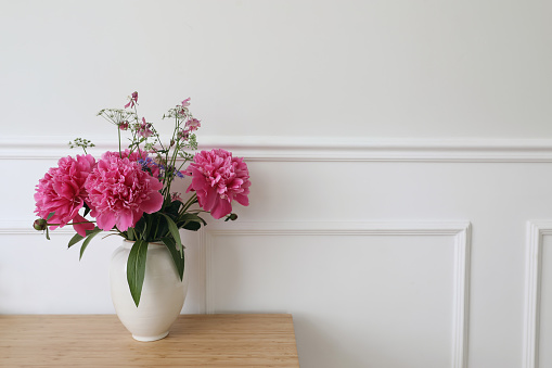 Beautiful floral bouquet. Vase with pink peonies flowers on ooden table, desk. White wall background with elegant moulding. Wedding or birthday concept, elegant interior still life, web banner.