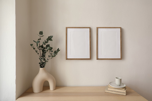 Two vertical picture frame mockup hanging on beige wall. Cup of coffee, books on wooden table. Vase with silver eucalyptus tree branches. Elegant interior, beautiful home decor, trendy working space.