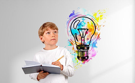 School kid taking notes in notebook, looking up at colorful light bulb doodle on white shadow wall. Concept of education, idea, plan and start up