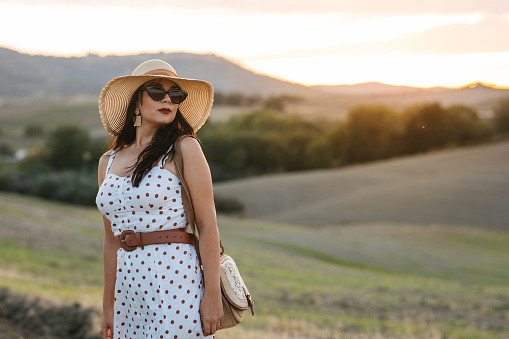 Portrait of a well-dressed woman with dress, hat and sunglasses posing in countryside of Italy. Visiting Tuscany in September