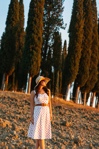 Portrait of a well-dressed woman with dress, hat and sunglasses posing in countryside of Italy at sunset. Visiting Tuscany in September. Cypress trees