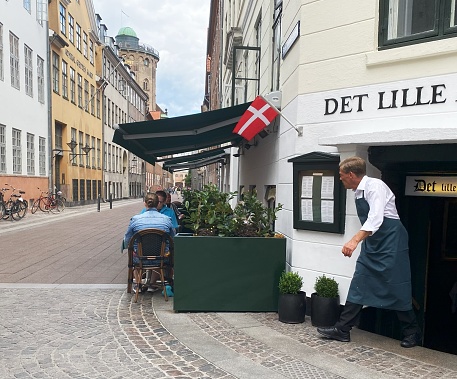 Waiter comes up the stairs from the restaurant to serve the customers sitting outdoors. The photo was taken in Store Kannike Stræde down town Copenhagen, Denmark on July  17th, 2023.