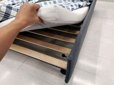A man's hand is lifting a mattress to expose the wooden slats used to support the mattress of the bed.