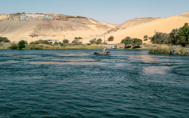 Aswan, Egypt - Feb 7, 2025:  Local Egyptians sailing a felucca on the Nile River at Aswan. First Cataract with large sand dunes in the background. Authentic cultural travel scene.