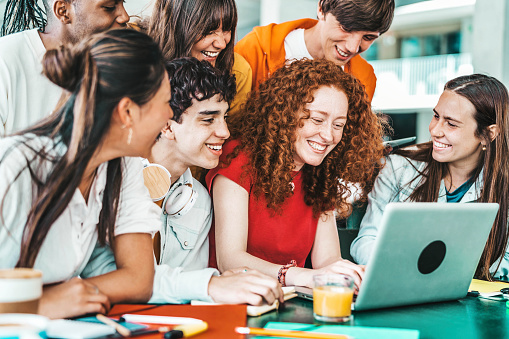 Multiracial university students sitting together at table with books and laptop - Happy young people doing group study in high school library - Life style concept with guys and girls in college campus