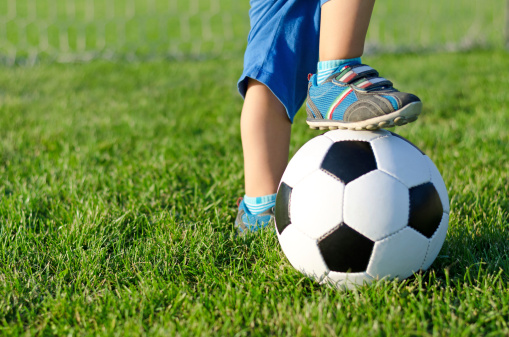 Little boy in shorts and trainers with his foot resting on top of a soccer ball on green grass with copyspace