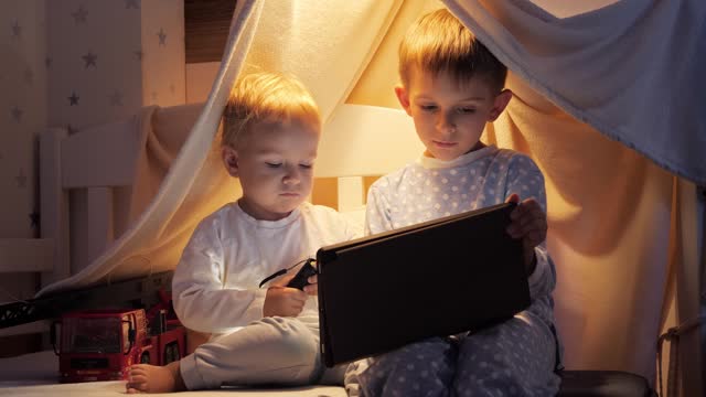 Two brothers in pajamas watching video on tablet computer in bed at night. Children with gadgets, education, kids development.