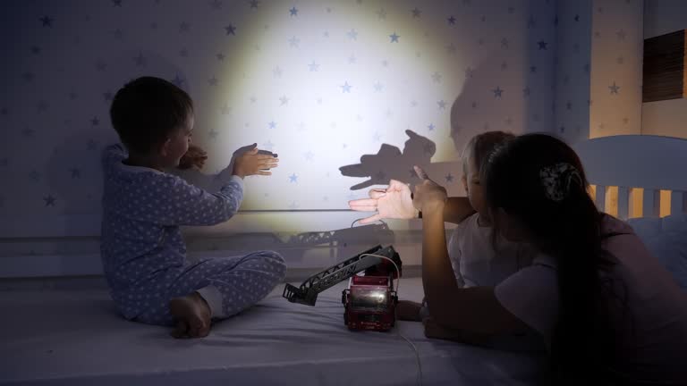 Young mother making shadow theater with her son wearing pajamas at night. Family having time together, parenting, happy childhood and entertainment