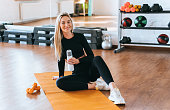 Cheerful blonde caucasian young woman in black sportswear sits on yoga mat with Bottle of water looks at camera at fitness class. Satisfied Swedish girl trains at  sport club, break time. Succes