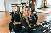 Blonde caucasian young woman in black sportswear and boxing gloves sitting at mirror at fitness club on step. Fitness, sport, real people. American fit girl training at sport club. European female