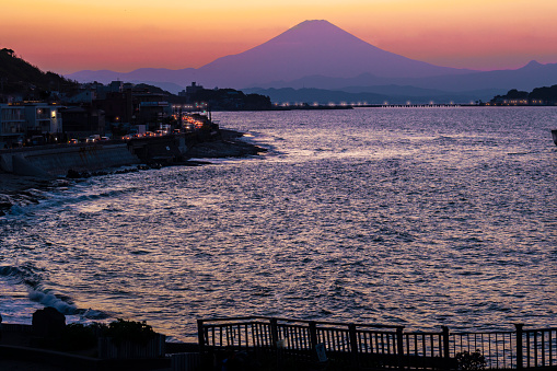 Enoshima Coast,Inamuragasaki, Kamakura, Kanagawa ,Japan-March 8 ,2023: Two people at the coast near Enoshima and Inamuragasaki beach in Fujisawa city, Kanagawa, Japan, marvel at the breathtaking view of Mount Fuji at dusk, as a flock of birds gracefully flies overhead.This captivating image features a serene, picturesque twilight scene shot near Enoshima and Inamuragasaki beach in Fujisawa city, Kanagawa prefecture, Japan, with the magnificent Mount Fuji as the focal point. As the sun dips behind the majestic mountain range, the vibrant hues of sunset paint the sky with ethereal beauty. This image perfectly encapsulates a profound sense of wonder and admiration for the natural world.