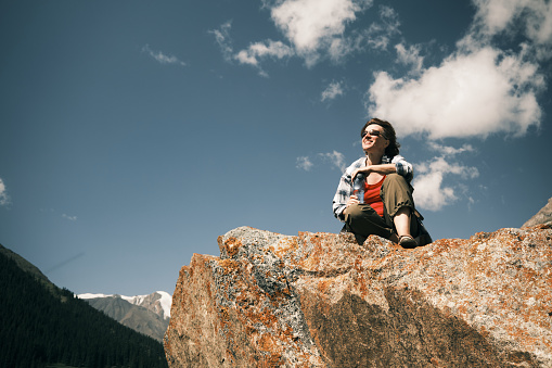 Cheerful 46-year-old female tourist is sitting on the rock high in mountains and drinking water