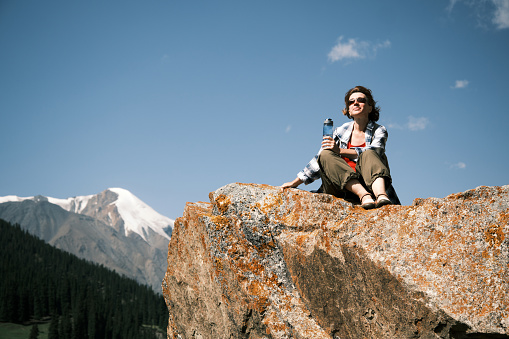Cheerful 46-year-old female tourist is sitting on the rock high in mountains and drinking water