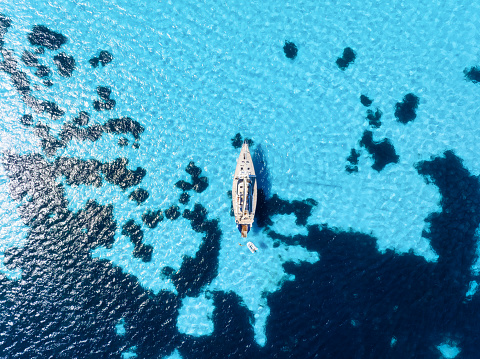 A yacht near the reef. Sail yacht on the sea as a background. 
Sea and waves from top view. Blue water background from top view. Top view from drone. Summertime vacation.